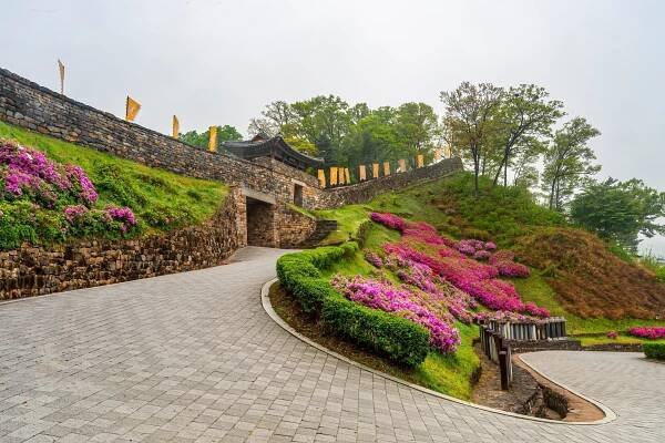 A winding cobblestone pathway leads to an ancient stone gateway adorned with traditional flags, surrounded by lush greenery and vibrant purple azalea bushes.