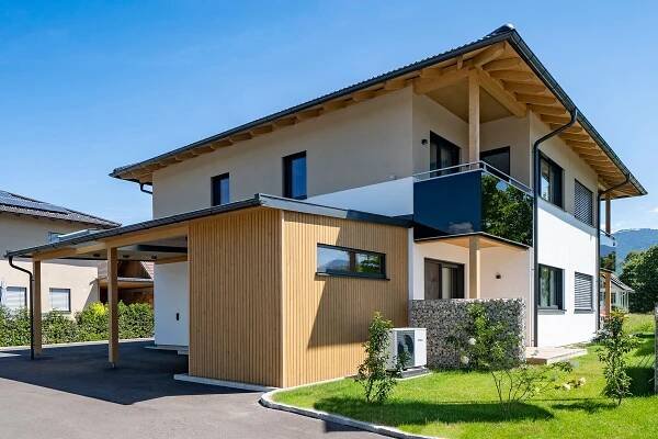 Modernes ökologisches Doppelhaus in Koblach 116 mit Holzverkleidung und Balkon, umgeben von gepflegtem Garten und blauem Himmel, ideal für nachhaltigen Hausbau.