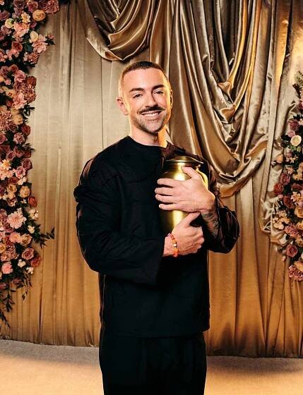 Matteo Lane holding an urn standing in front of a decorative backdrop with flowers and curtains