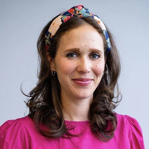 A woman with long brown hair wearing a colorful floral headband and a bright pink top, smiling gently in front of a plain light gray background.