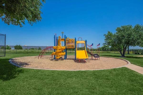 A brightly colored playground featuring a yellow slide, climbing structure, and various play equipment, surrounded by grass and trees. There is a gravel area beneath the playground, and a paved path leading toward it. In the background, multiple flags are visible against a clear blue sky.
