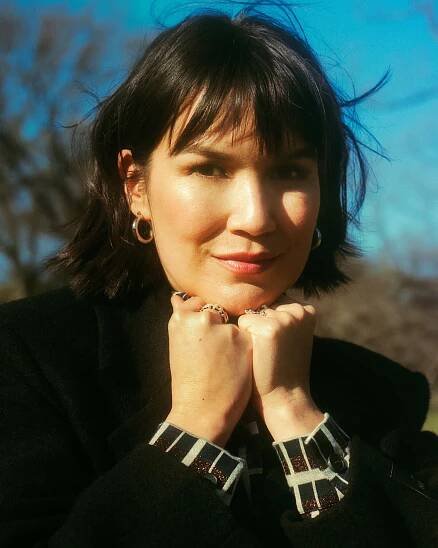 Zoë Chao, in a black coat and wearing chunky jewelry, rests her chin on her fists outdoors and looks into the camera. 