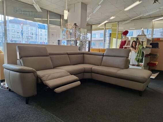 Šedá sekční pohovka ve tvaru písmene L s jedním polohovatelným sedadlem je vystavena v showroomu nábytku s velkými okny, horním osvětlením a plakátovým stojanem v pozadí.