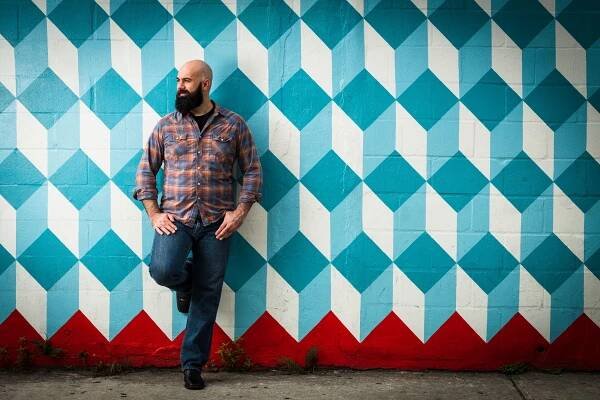 A portrait of a bald man with a beard wearing a plaid shirt and jeans leaning against a mural of three dimensional blue squares.
