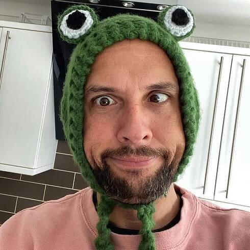 Mike Jordan wearing a green knitted bonnet that has knitted eyes to look like a frog. He is pulling a silly face