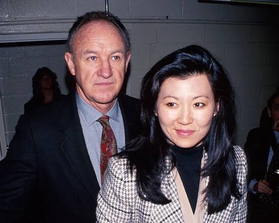 Betsy Arakawa stands in front of Gene Hackman. He is dressed in a suit; she has her hair down and is wearing a blazer with shoulder pads.