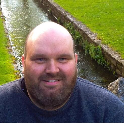 Photo of a man with a beard, smiling, near a stream.