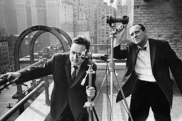 A black and white photo of two men in suits peering into telescope-like devices positioned on a building terrace high above city streets.