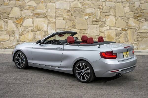 Silbernes Cabriolet mit roter Innenausstattung vor einer Steinmauer, aus der Hecksicht aufgenommen. Fahrzeug hat ein offenes Verdeck.