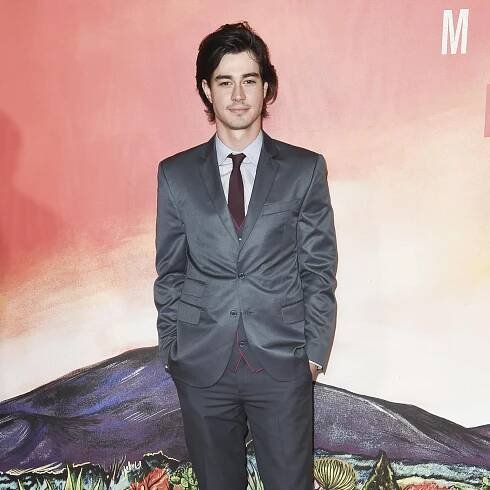 Man with long black hair in a grey suit wears a white collared shirt and a red tie as he poses for photos on a red carpet