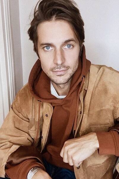 lucas bravo wearing a rust colored hoodie and tan suede jacket looking at the camera