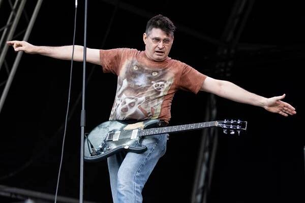 A man with arms outstretched, in glasses, a brown T-shirt with animals on it and jeans with a guitar onstage.