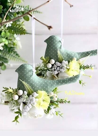 two green birds hanging from a branch with flowers and greenery on it's sides