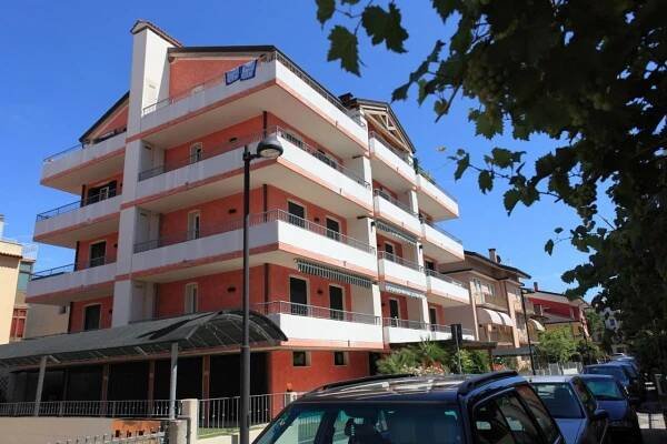 Pětipatrová červeno-bílá budova s balkony a zaparkovanými auty před ní.