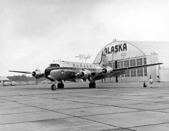 Propeller plane on a runway with an airplane hanger behind it, and Alaska spelled out in large blocky letters. 