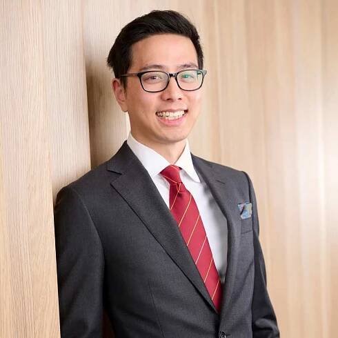 Link to William Li Profile Page. A man wearing glasses and a dark suit with a red striped tie leans against a wooden wall, smiling. The background is a light wooden texture, and the man has short dark hair. He appears confident and approachable.