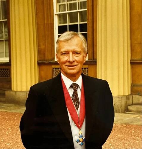 A man with white hair in a dark suit, white shirt and patterned tie is wearing a red ribbon with a blue and gold cross hanging from it. He has just been awarded a CBE