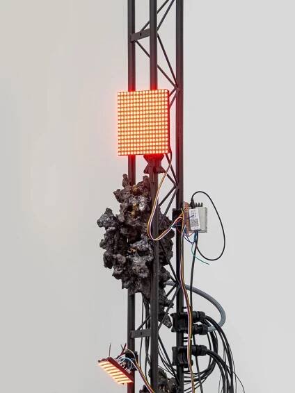 A close-up of a metallic structure featuring red LED lights and tangled wires, alongside a textured, rocky material.
