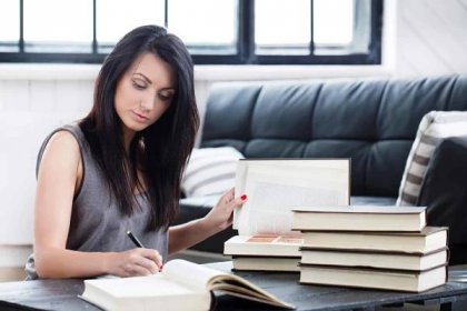 A young woman with black hair sitting down on the floor with a stack of books on the table in front of her. She is holding a book open with her left hand, and writing something with a pen in her right hand in another book. A black sofa and window is seen in the background.