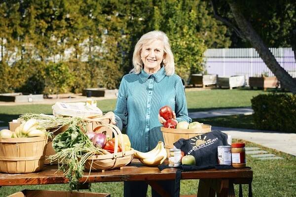 Photo: A white woman with a teal button up and teal slacks poses for a photo. She smiles for the photo with her blonde hair. She poses with her products on a table in front of her.