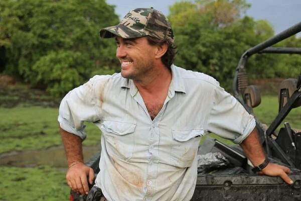 A man in work clothes, wearing a cap and leaning on against an off-road vehicle.