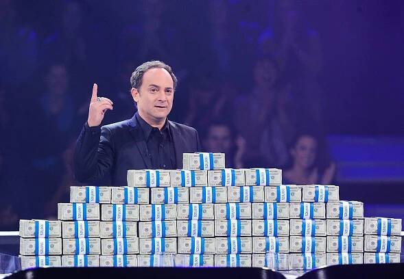 Kevin Pollak standing behind piles of cash