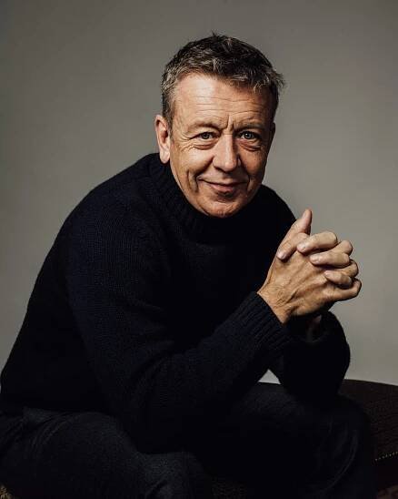 A portrait of Peter Morgan, with his hands clasped, wearing a chunky dark sweater.