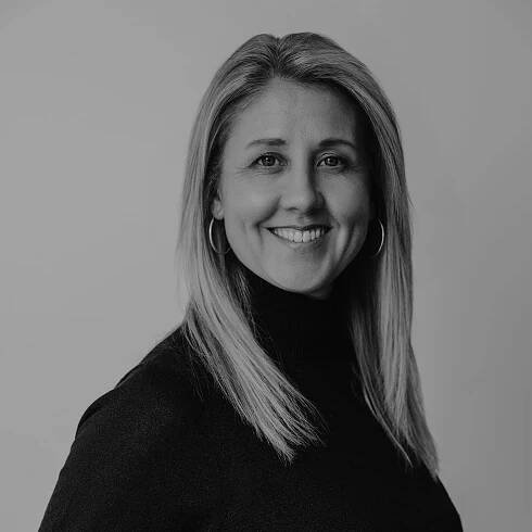 Black and white portrait of Rachel Bower, a woman with straight, shoulder-length hair, wearing hoop earrings and a long-sleeve turtleneck sweater, smiling while looking slightly off camera. Plain background.