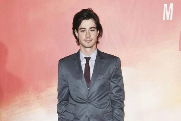 Man with long black hair in a grey suit wears a white collared shirt and a red tie as he poses for photos on a red carpet