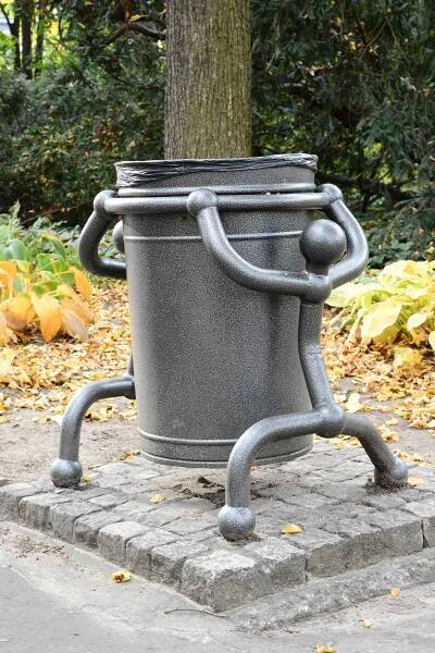 a metal trash can sitting on top of a sidewalk next to a tree and bushes