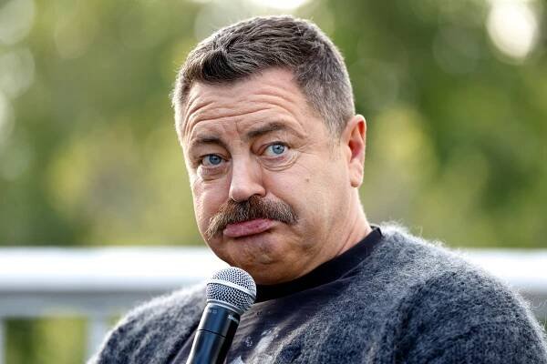 Man with a mustache and short hair holding a microphone, wearing a fuzzy sweater, looking amused at an outdoor event