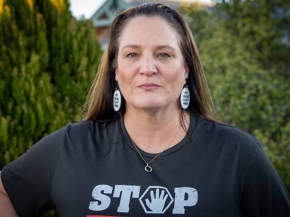 A woman has long brown hair, she looks solemn. She wears earrings which read "stop violence against women"