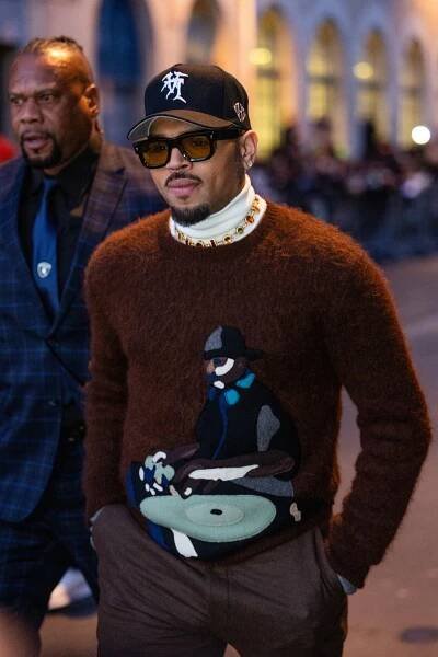 A man in a baseball cap, sunglasses and a jeweled necklace walks along a Paris street.