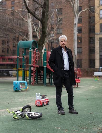 Jonathan Haidt, wearing a dark coat over a light shirt and dark pants, stands in a playground, with a bicycle and toys strewn on the ground.