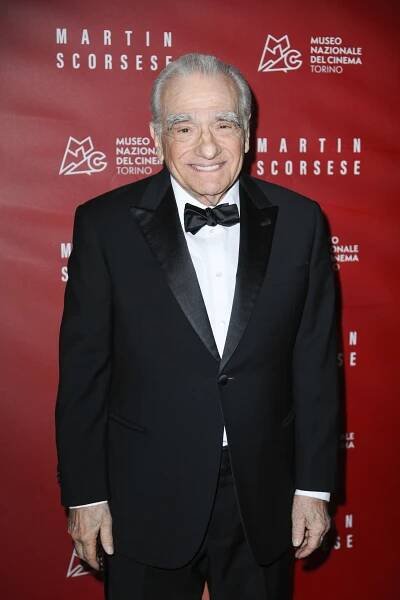 An unidentified man in a classic black tuxedo and bow tie stands smiling on a red carpet at an event with a backdrop featuring text and logos