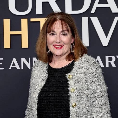Person smiling at an event, wearing a textured jacket over a black top, in front of a 'Saturday Night Live' anniversary backdrop