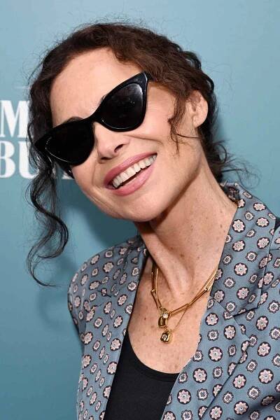 Minnie Driver with a low curly bun while wearing sunglasses and a blue-gray blazer