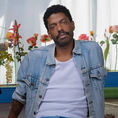artist Devan Shimoyama wearing a denim jacket over a white tank while he sits on the floor in front of flowers