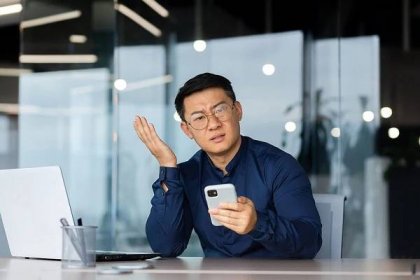 nespokojený asiat se dívá na fotoaparát, muž v kanceláři držící telefon, portrét nespokojeného podnikatele na pracovišti u přenosného počítače - notebook použití počítače fotky - stock snímky, obrázky a fotky