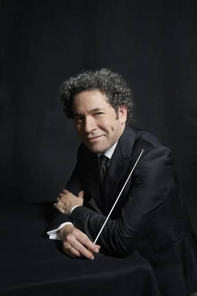 Gustavo Dudamel mit lockigem Haar im Anzug lehnt an einem dunklen Tisch und hält einen Taktstock vor einem dunklen Hintergrund. Er hat Grübchen und blickt direkt in die Kamera.