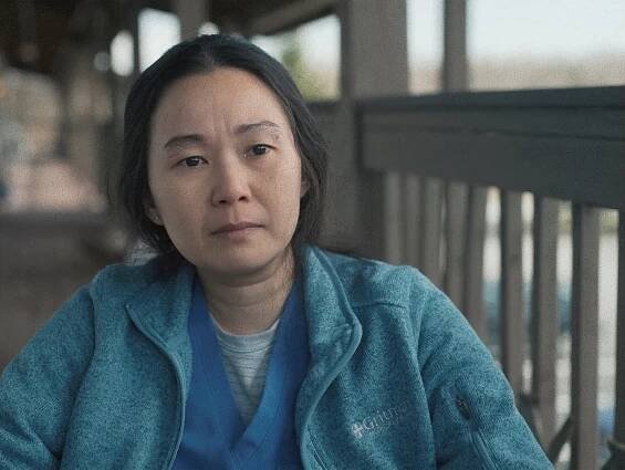In a scene from the film, Chau is facing the camera in a gray outdoor walkway. She’s wearing a teal zip-up jacket over blue nurse’s scrubs and gazing sadly into the distance.