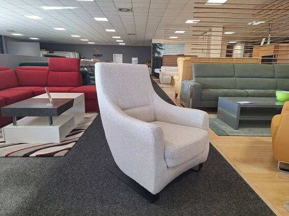 V moderním showroomu nábytku jsou vystaveny pohovky v různých barvách. Na tmavém koberci uprostřed je výrazně umístěno světle šedé čalouněné křeslo, které je obklopeno červenými, zelenými a béžovými pohovkami.