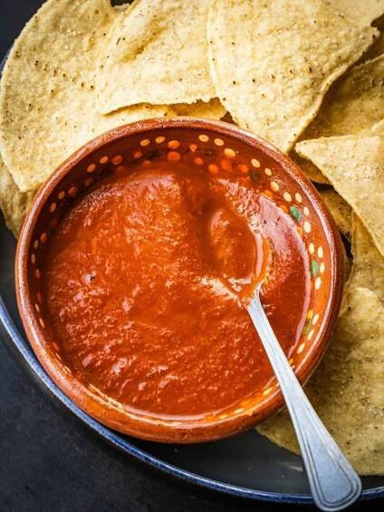 Salsa roja in a terracotta bowl with a spoon on a plate with some tortilla chips.