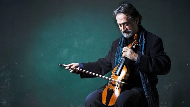 Jordi Savall mit grauem Haar und Bart sitzt vor einem dunkelgrünen Hintergrund, hält eine Gambe und einen Bogen und trägt einen dunklen Anzug mit einem blauen Schal.