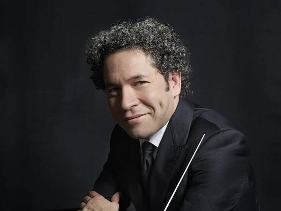 Gustavo Dudamel with curly hair in a suit leans against a dark table and holds a baton in front of a dark background. He has dimples and looks directly into the camera.