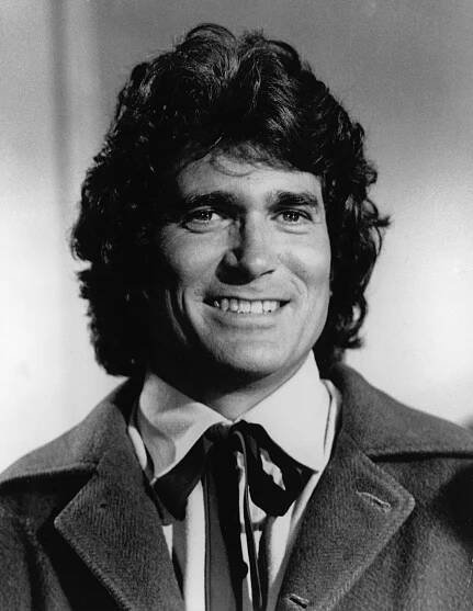A person with wavy hair wearing a collared shirt and jacket smiles warmly at the camera. 
