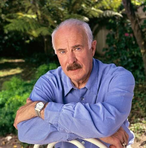 A portrait of Dabney Coleman, a man with thinning white hair and a dark mustache, sitting outdoors in a chair with his arms crossed and wearing a dark blue shirt.