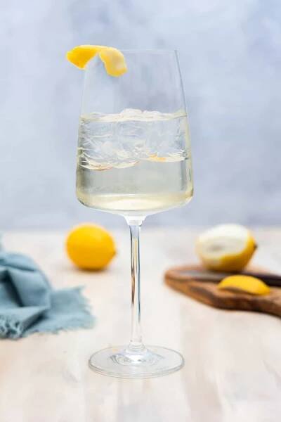 A wine glass filled with ice and an almost clear liquid, with a lemon twist garnish on the left side of the glass.