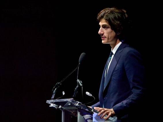 Jack Schlossberg in a suit speaks at a podium with microphones, set against a dark background