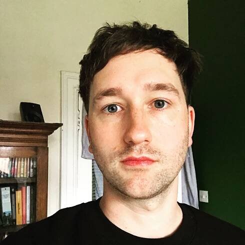 Headshot of author, Charlie Lynch, a white male with dark hair and green eyes, with some stubble.